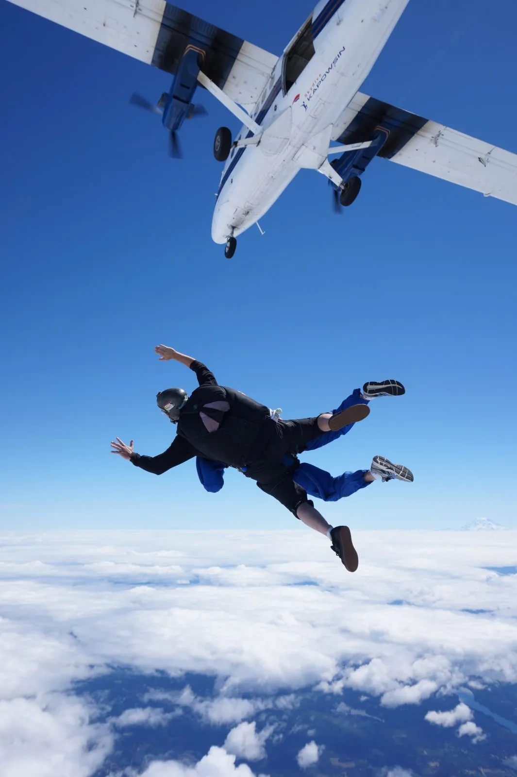 A skydiver leaping from the plane into open blue sky, high above a blanket of clouds.